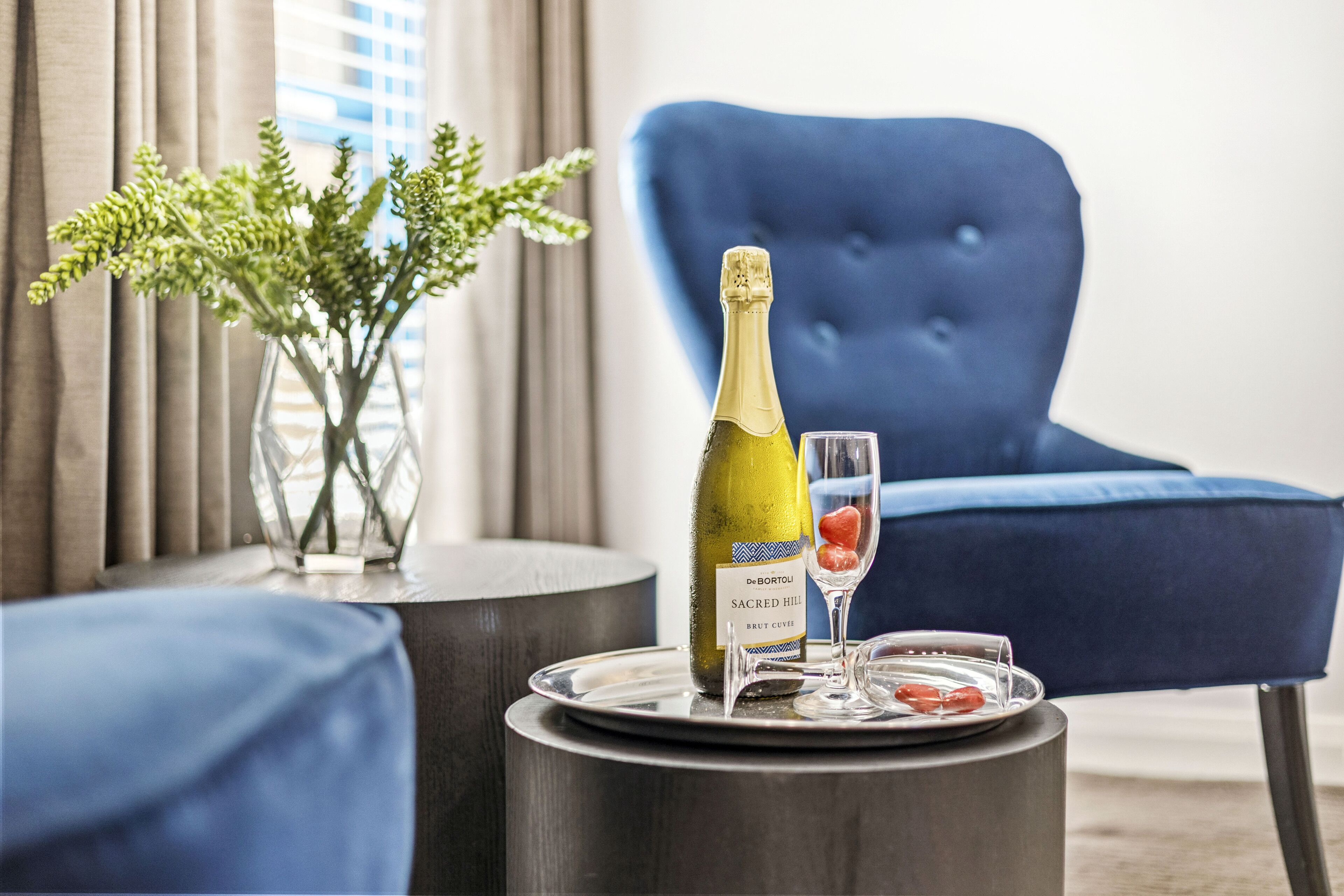 A bottle of wine on a tray with a glass, a vase with green plants, and a blue chair in a room.