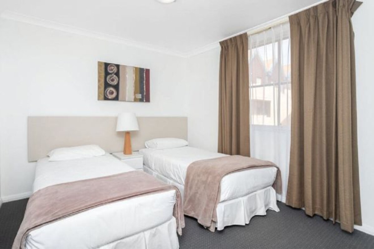 Habitación de hotel con dos camas, una mesita de noche, una lámpara, un cuadro en la pared y una ventana con cortinas.
