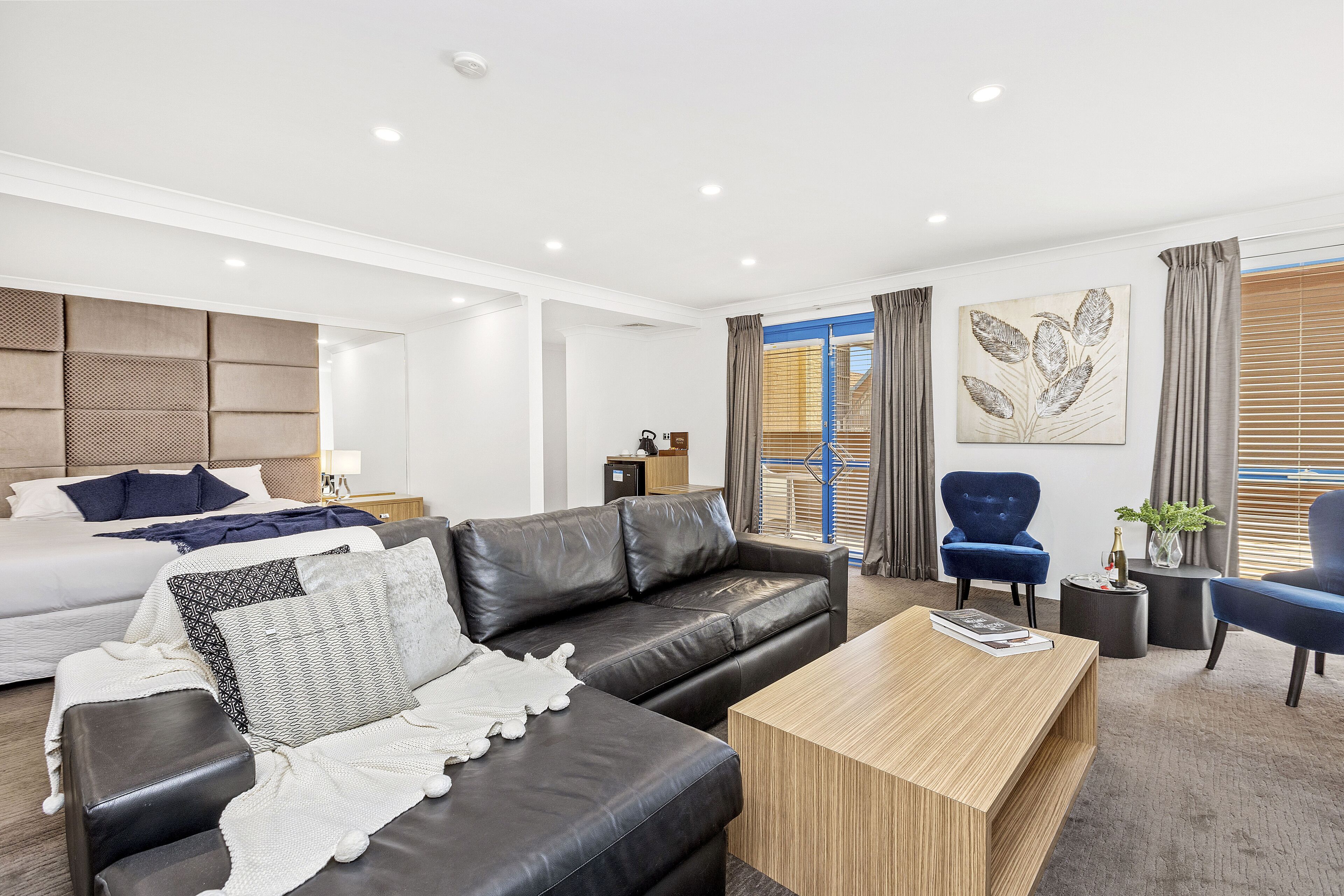 A modern hotel room with a large bed, a black leather sofa, a wooden coffee table, and a blue armchair.