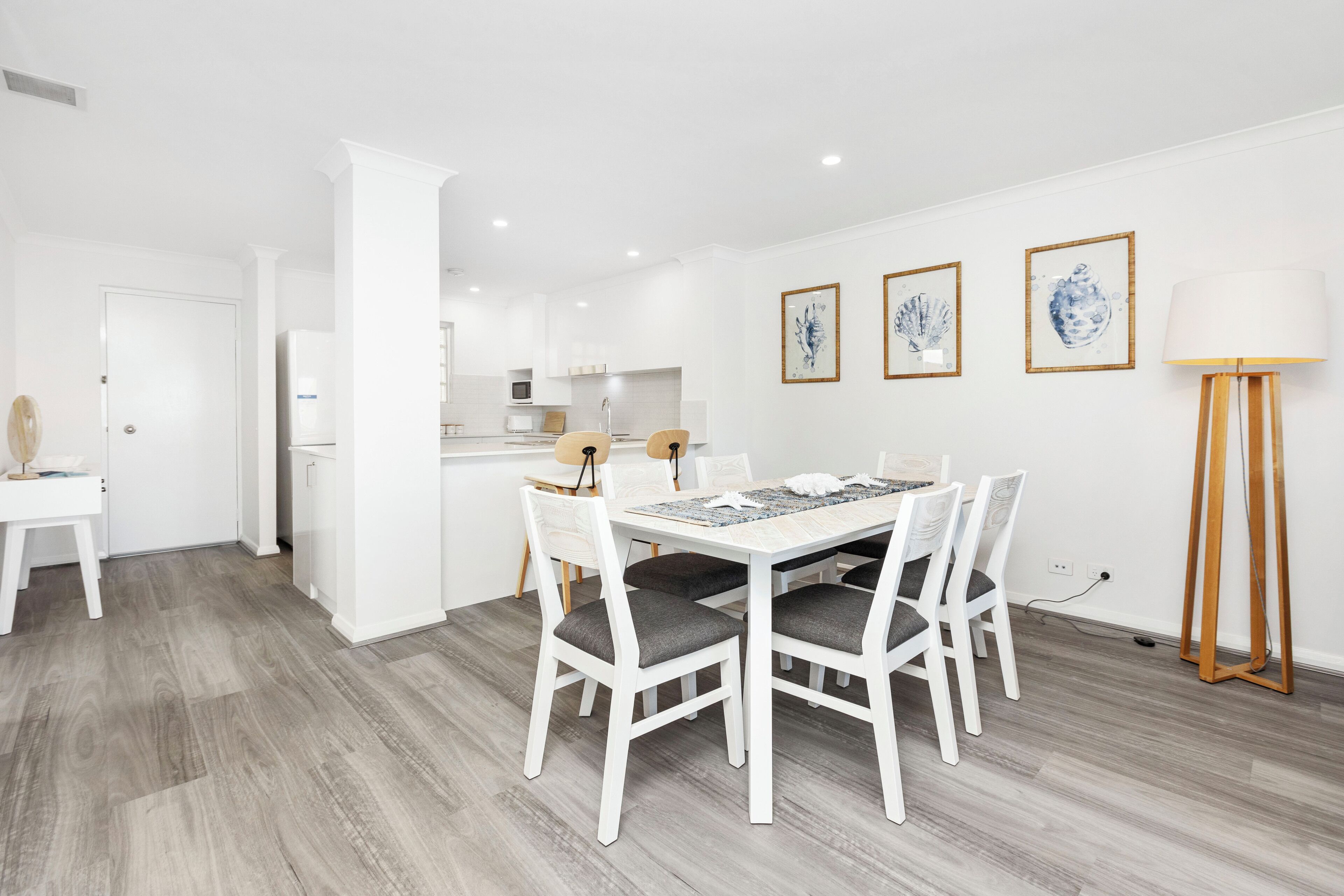 Ett modernt kök med vita skåp, en matplats med ett vitt bord och stolar samt tre inramade konstverk på väggen.
