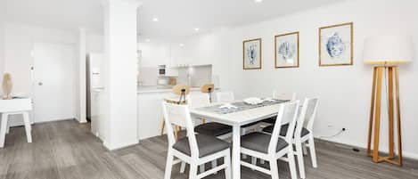 A modern kitchen with white cabinets, a dining area with a white table and chairs, and three framed artworks on the wall.