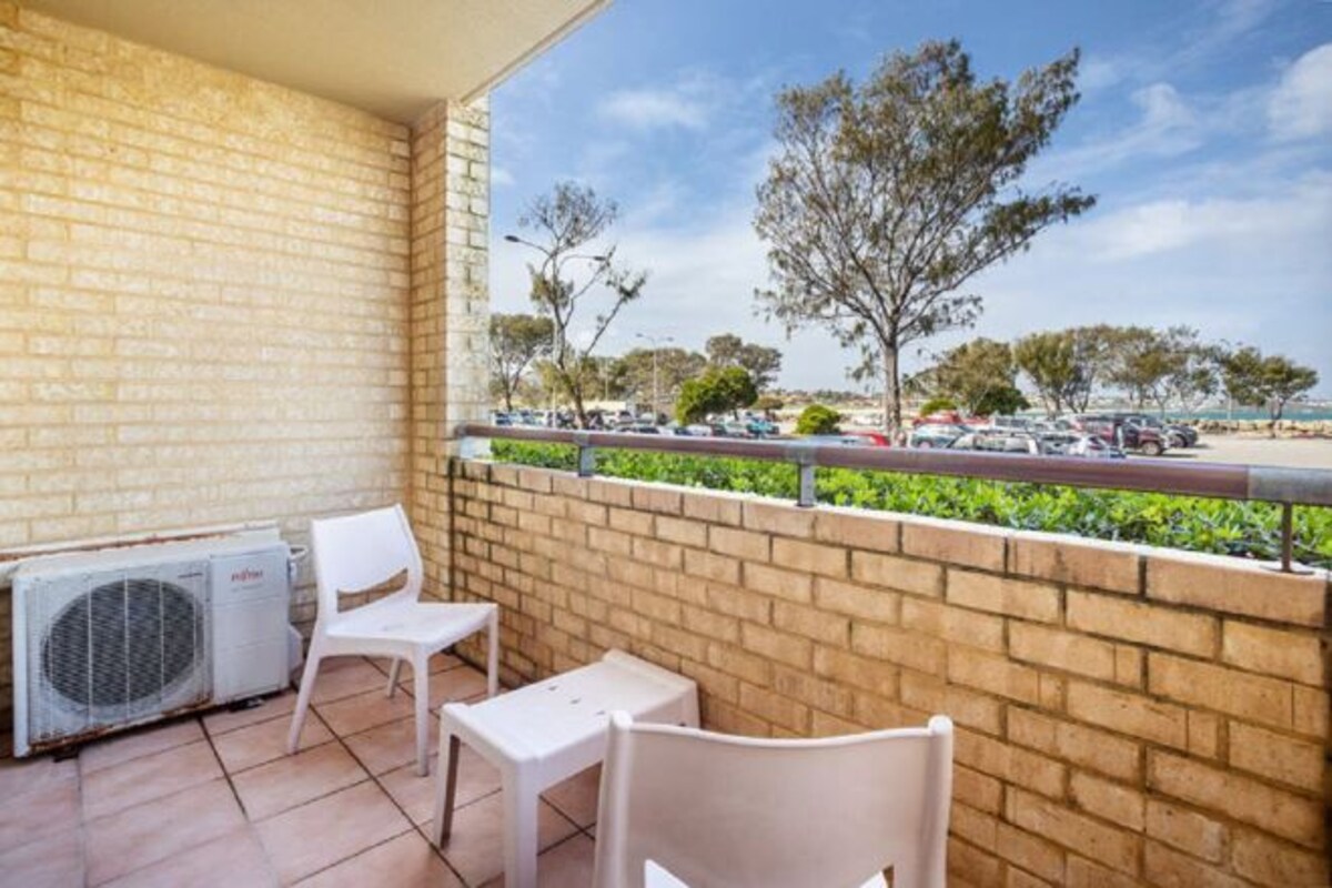 Un balcón con pared de ladrillo, una silla blanca, una mesita y un equipo de aire acondicionado.