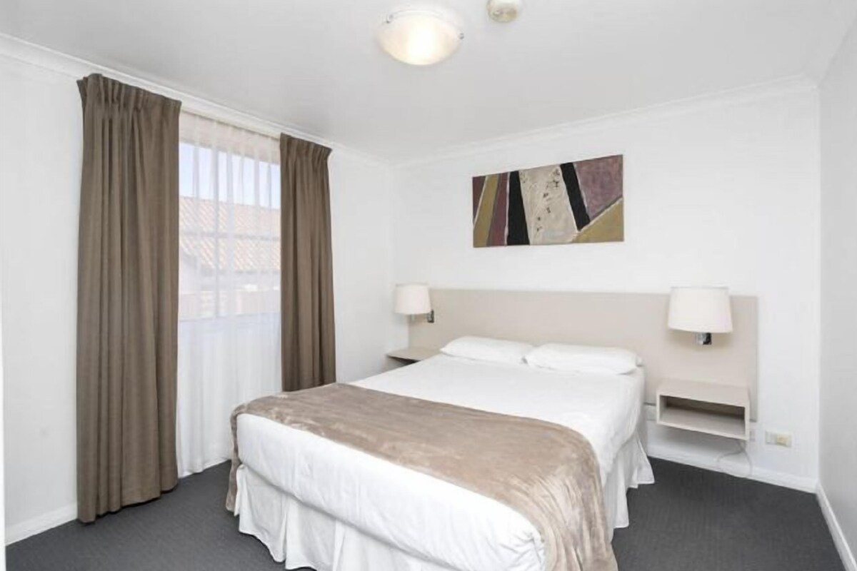 A hotel room with a large bed, two bedside tables, a wall-mounted TV, and a window with curtains.