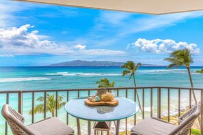 A balcony with a glass table and wicker chairs, overlooking a turquoise ocean and palm trees. - Valley Isle Resort #707 - Vacation West Maui - Beachfront Luxury (Lahaina)