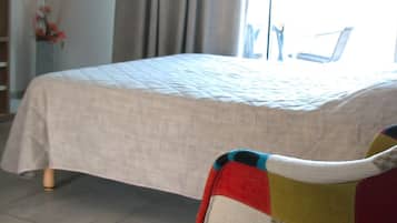 Un lit recouvert d’une simple couvre-lit, une chaise agrémentée d’un coussin coloré et une étagère en bois dans une pièce dont le sol est carrelé.