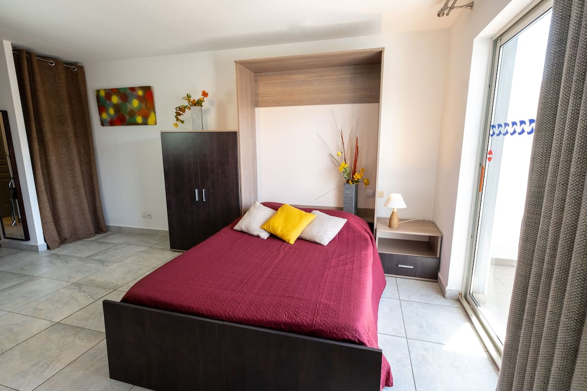 Une chambre moderne avec un grand lit, une armoire sombre, une petite table avec une lampe et une fenêtre avec des rideaux.