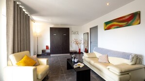 Uma sala de estar moderna com sofá, poltrona e mesa de centro. Há uma porta ao fundo e um quadro na parede.