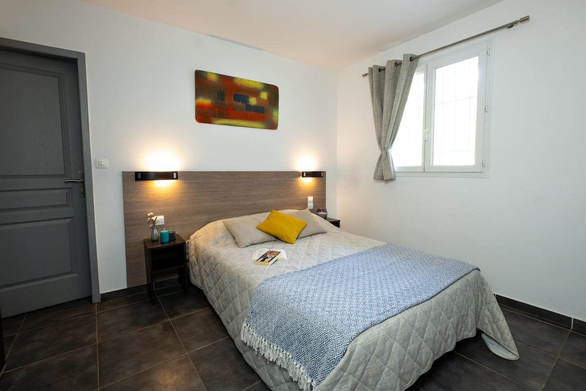 Une chambre à coucher comprenant un lit, des tables de chevet, une commode, une lampe fixée au mur, une fenêtre avec des rideaux et une porte.