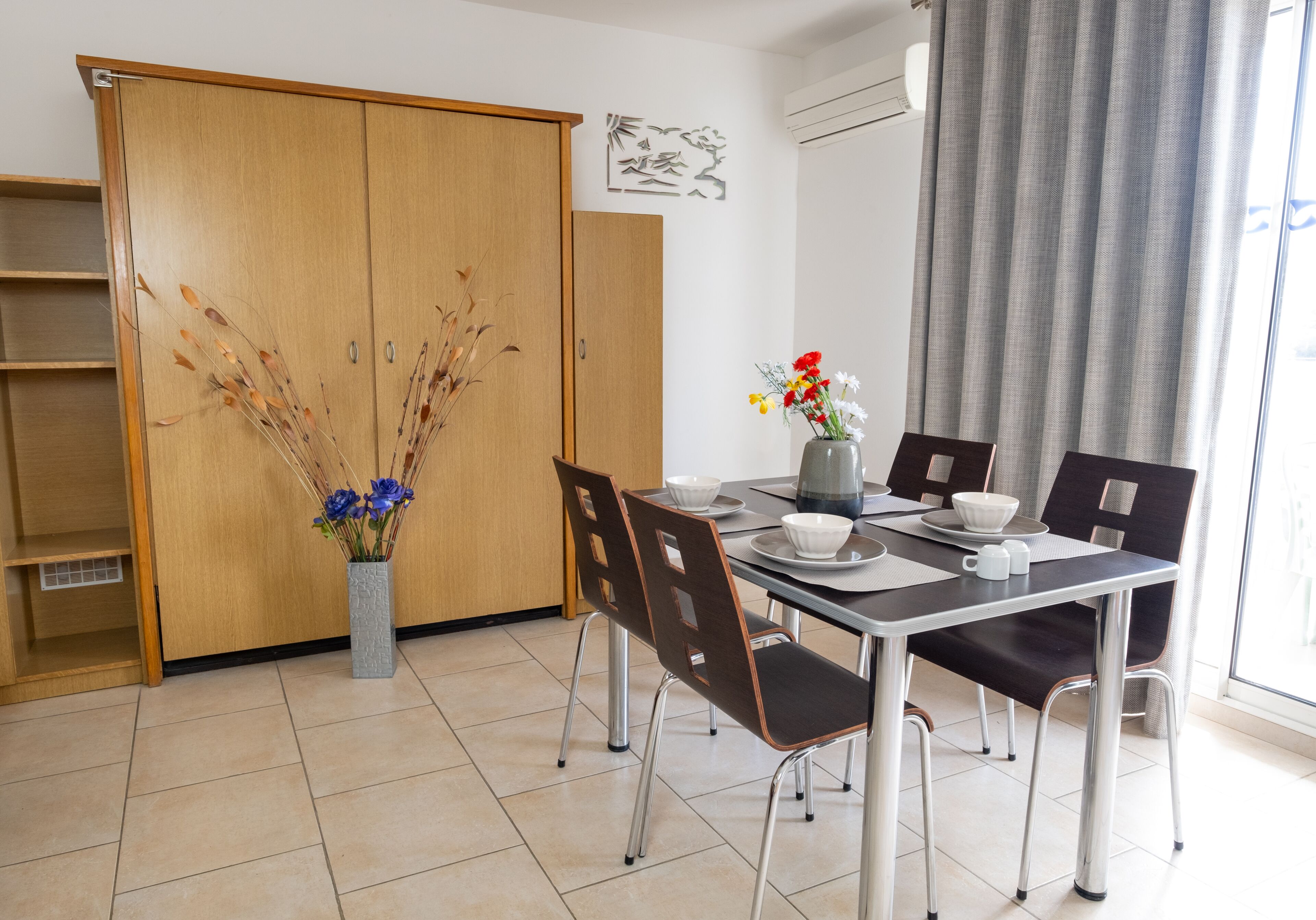 A dining area with a table set for four, surrounded by chairs, a vase with flowers, and a wooden wardrobe.