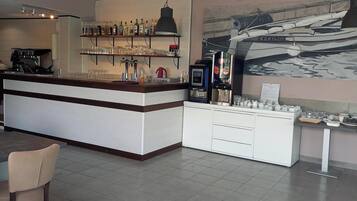 Zona de bar moderna con mostrador, estanterías llenas de botellas, vasos y una máquina de café. Hay una zona de estar con sillas y una mesa.