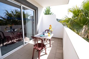 A balcony with a table and chairs, a vase with flowers, and a view of palm trees and a building. - Residence le Saint Esteve (La Ciotat)
