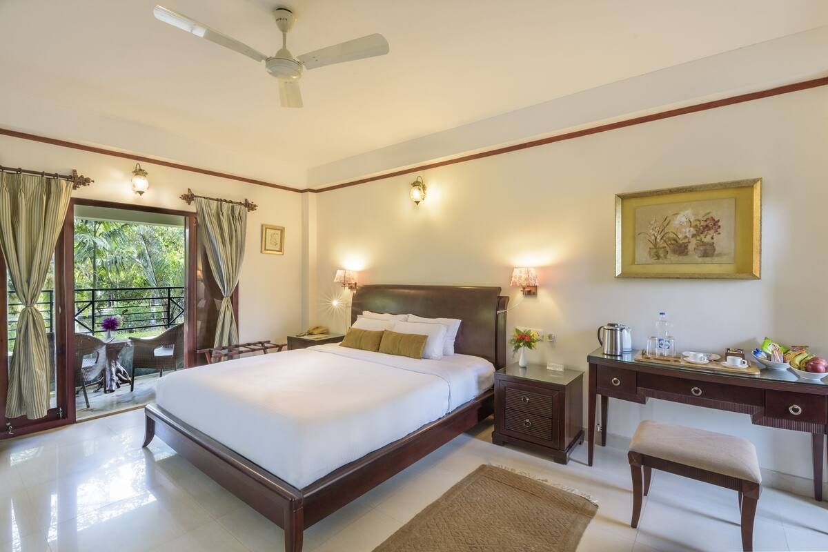 Habitación de hotel con una cama grande, un escritorio con una silla, una mesita de noche y vistas a zonas verdes a través de la puerta.