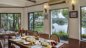 A dining area with a table set for breakfast, large windows with a view of a lake and greenery, and a wall-mounted air conditioner. - The Riverwood Forest Retreat - Dooars (Mal)