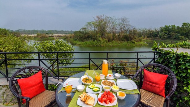A table set with breakfast items, including toast, watermelon, and a pitcher of orange juice, overlooking a river with greenery and trees. - The Riverwood Forest Retreat - Dooars (Mal)