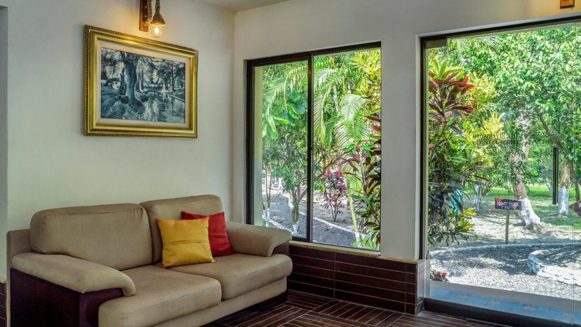 A room with a beige sofa, a framed picture on the wall, and large windows overlooking a garden.