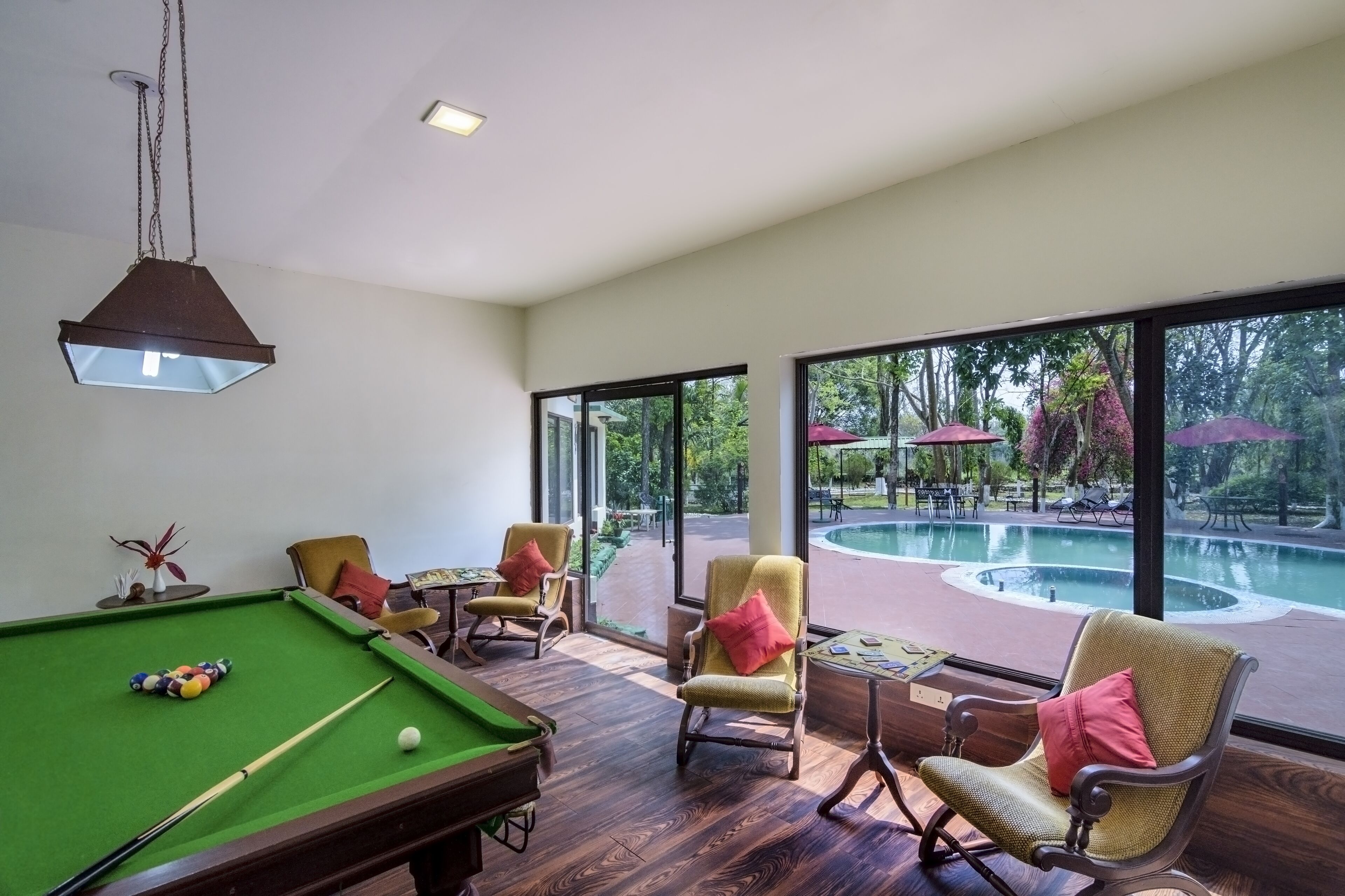 A pool table with pool balls, a billiard cue, and a green felt surface. There are two chairs with cushions, a small table with a card game, and a hanging light fixture. Large windows provide a view of an outdoor swimming pool area with umbrellas and greenery.
