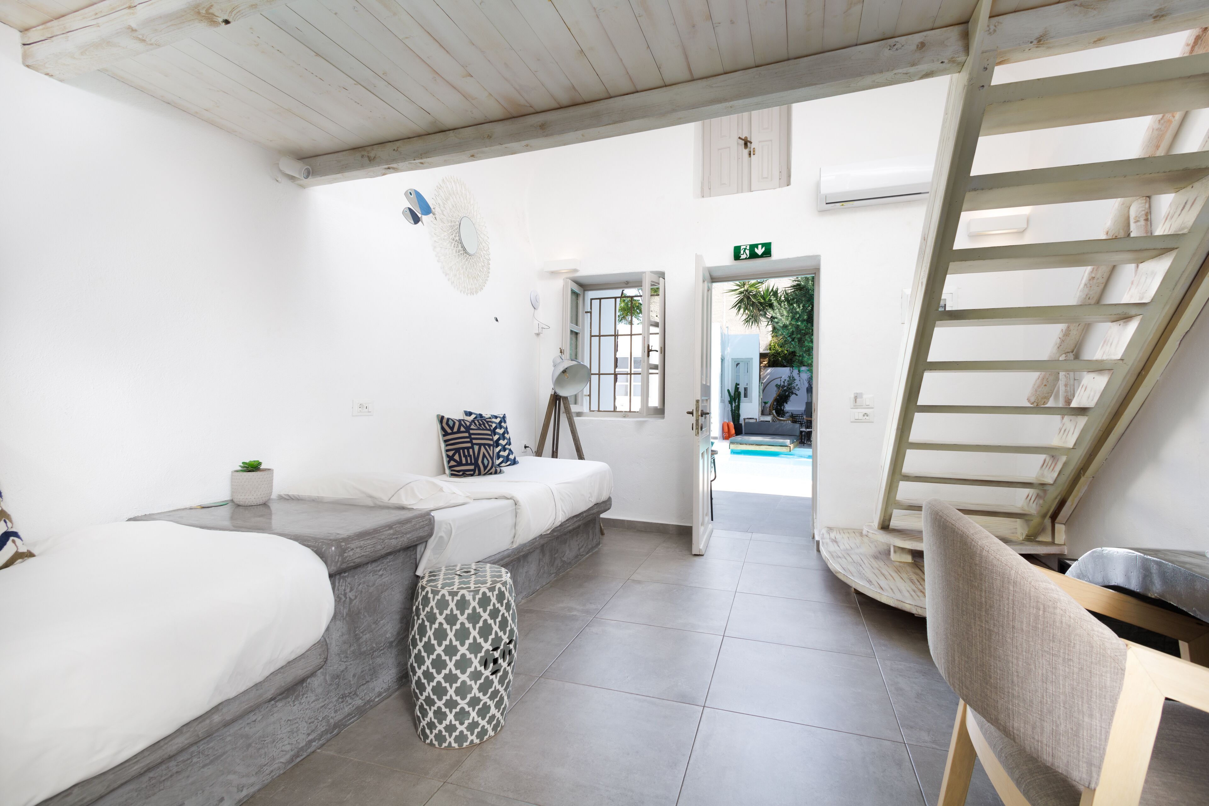 A modern, minimalist room with a bed, a small stool, and a chair. There is a staircase leading to an outdoor area.