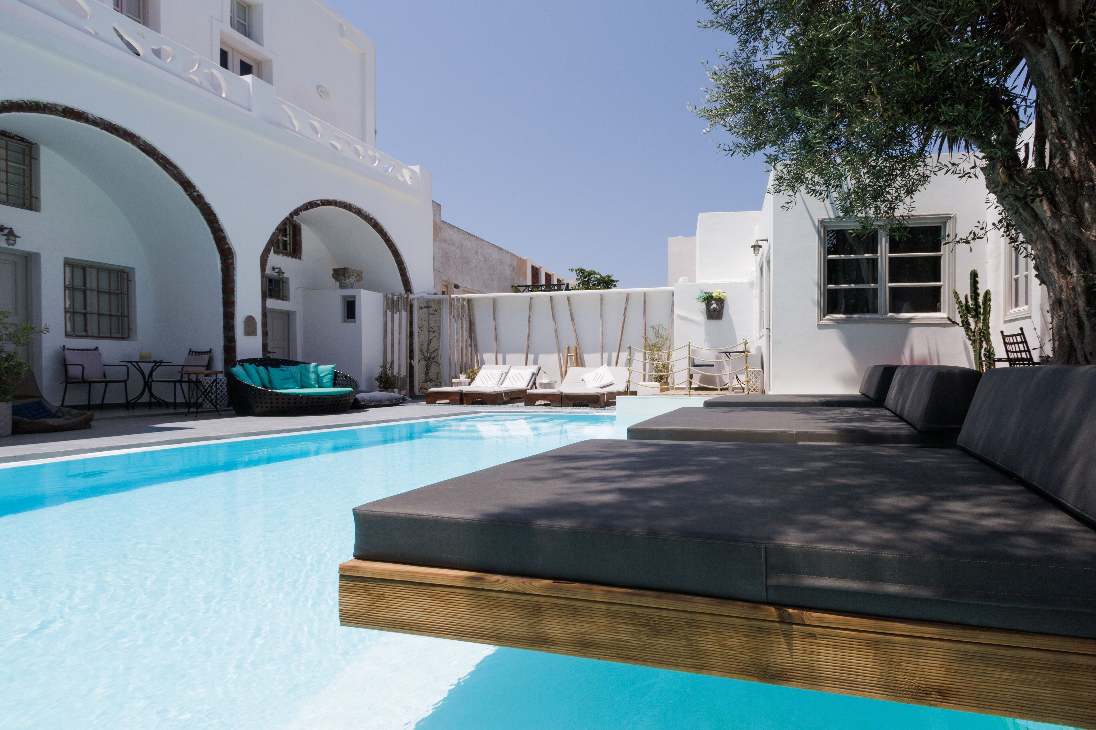 A swimming pool with lounge chairs and a sofa, surrounded by white buildings with arches.