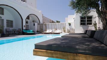 A swimming pool with lounge chairs and a sofa, surrounded by white buildings with arches.