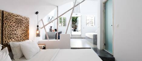 A modern hotel room with a large bed, a wooden headboard, a desk, and a view of the bathroom and outdoor area.