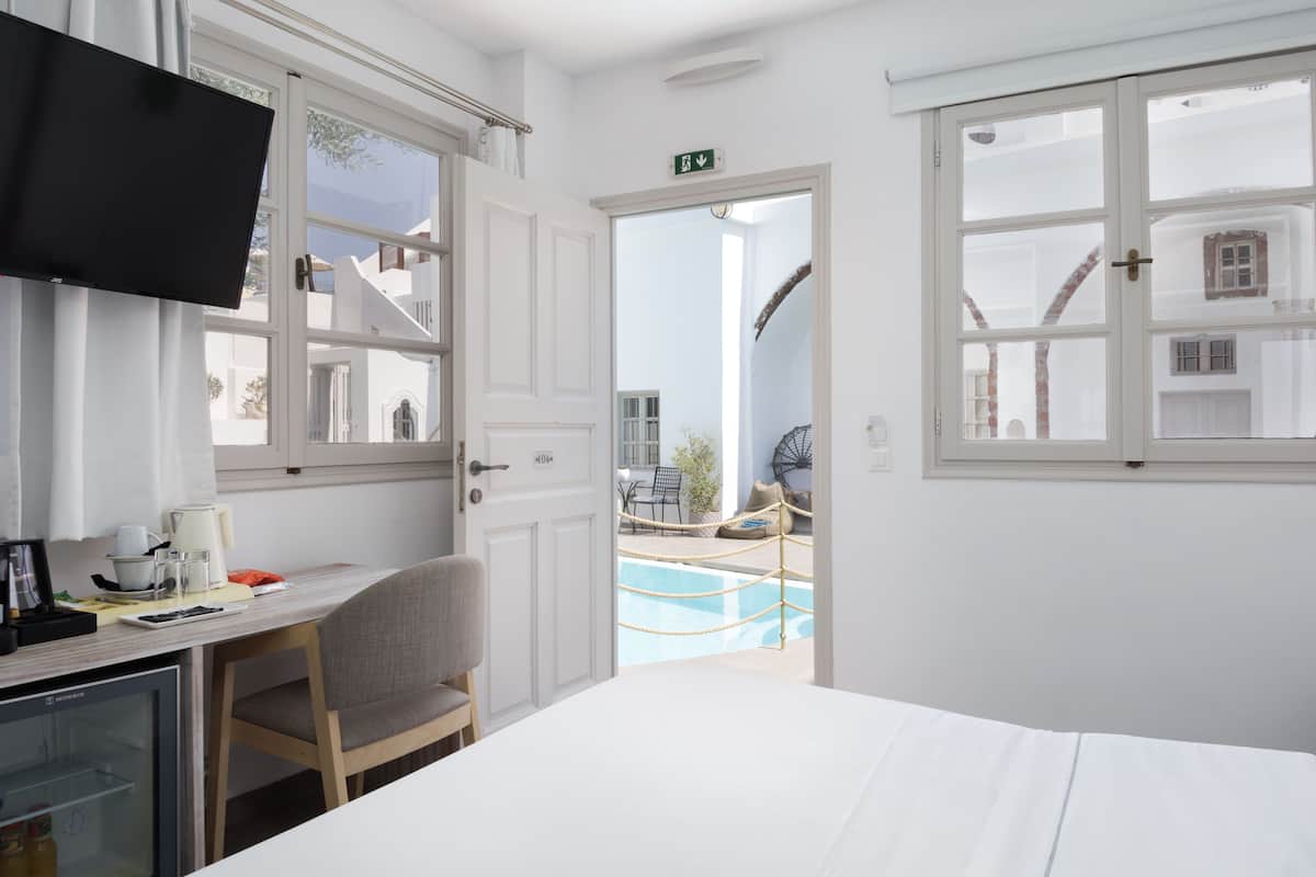 A hotel room with a bed, a desk with a chair, a TV, and a view of a pool and outdoor seating area.