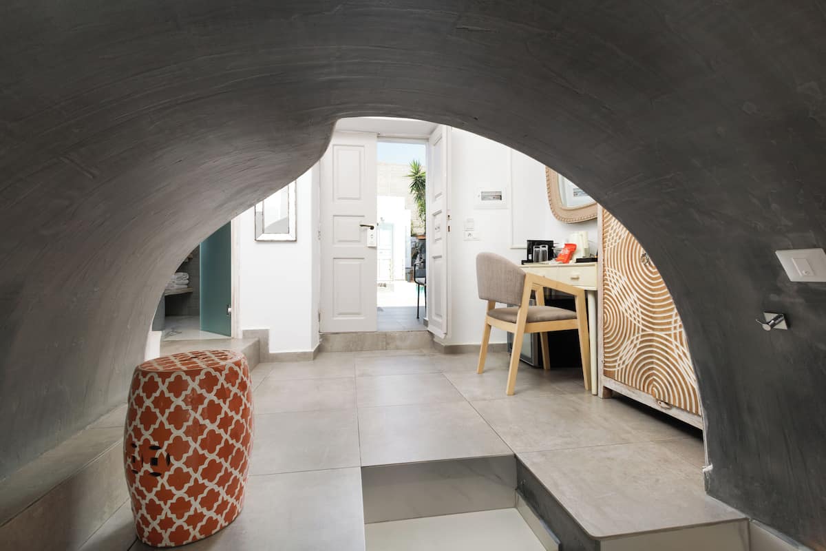 An interior space with a curved ceiling, a patterned ottoman, a dining area with a chair, and a kitchen visible through an open door.