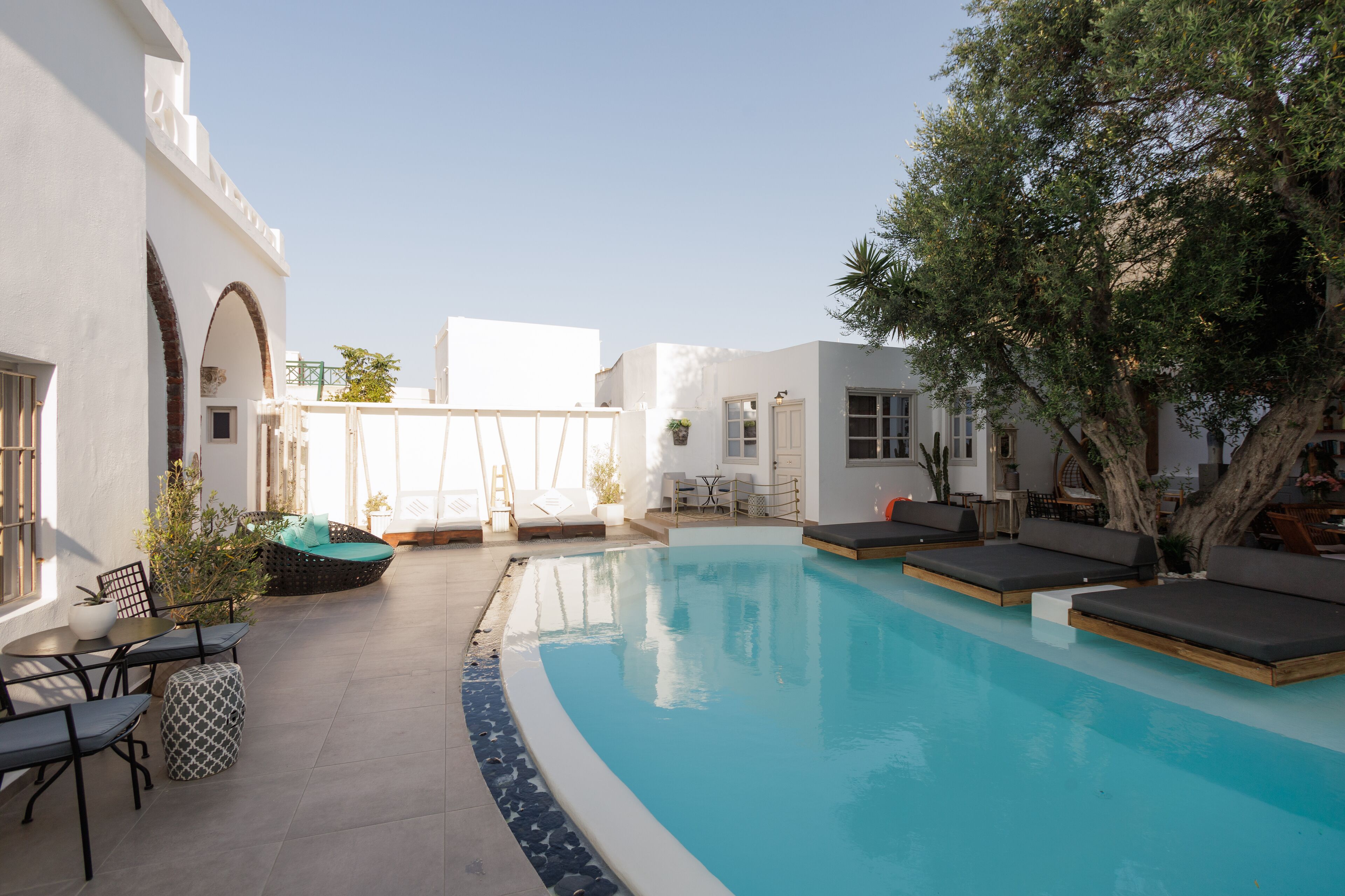 Zona de piscina con sillones de descanso y una mesa, rodeada de edificios blancos y un árbol.