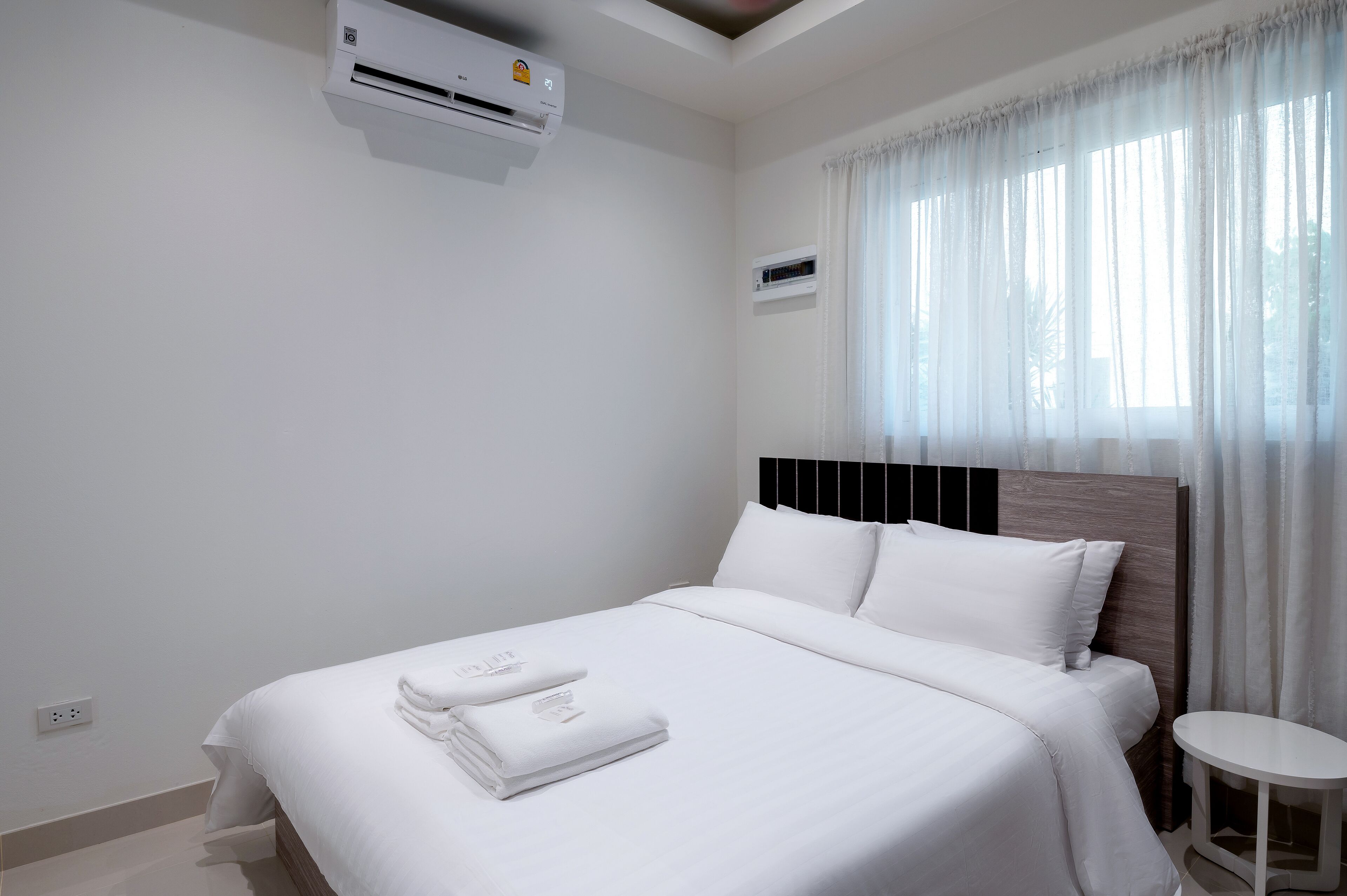 Una cama bien hecha con ropa blanca y una toalla doblada, una mesita de noche pequeña y una ventana con cortinas traslúcidas.