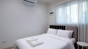 Una cama bien hecha con ropa blanca y una toalla doblada, una mesita de noche pequeña y una ventana con cortinas traslúcidas.