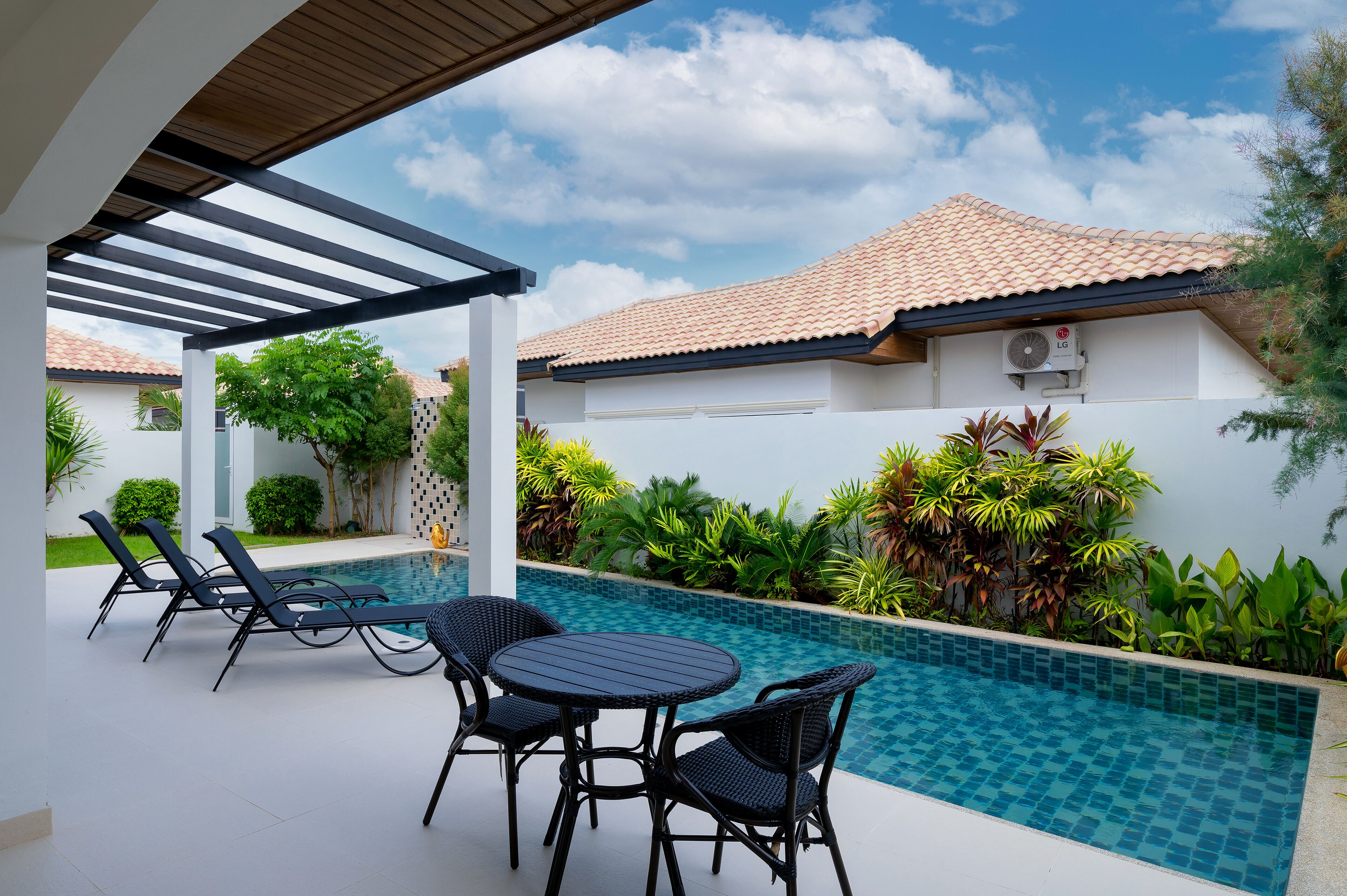 A pool area with lounge chairs and a table, surrounded by lush greenery and a white building with a tiled roof.