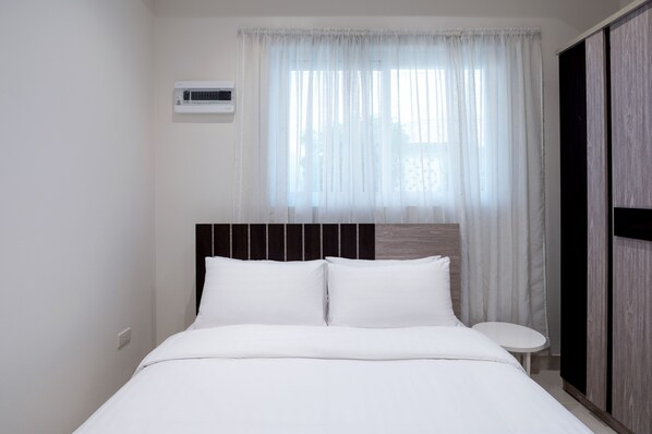 A neatly made bed with white linens and a headboard with vertical wooden slats. There is a small bedside table with a white surface and a wall-mounted air conditioning unit above the bed.