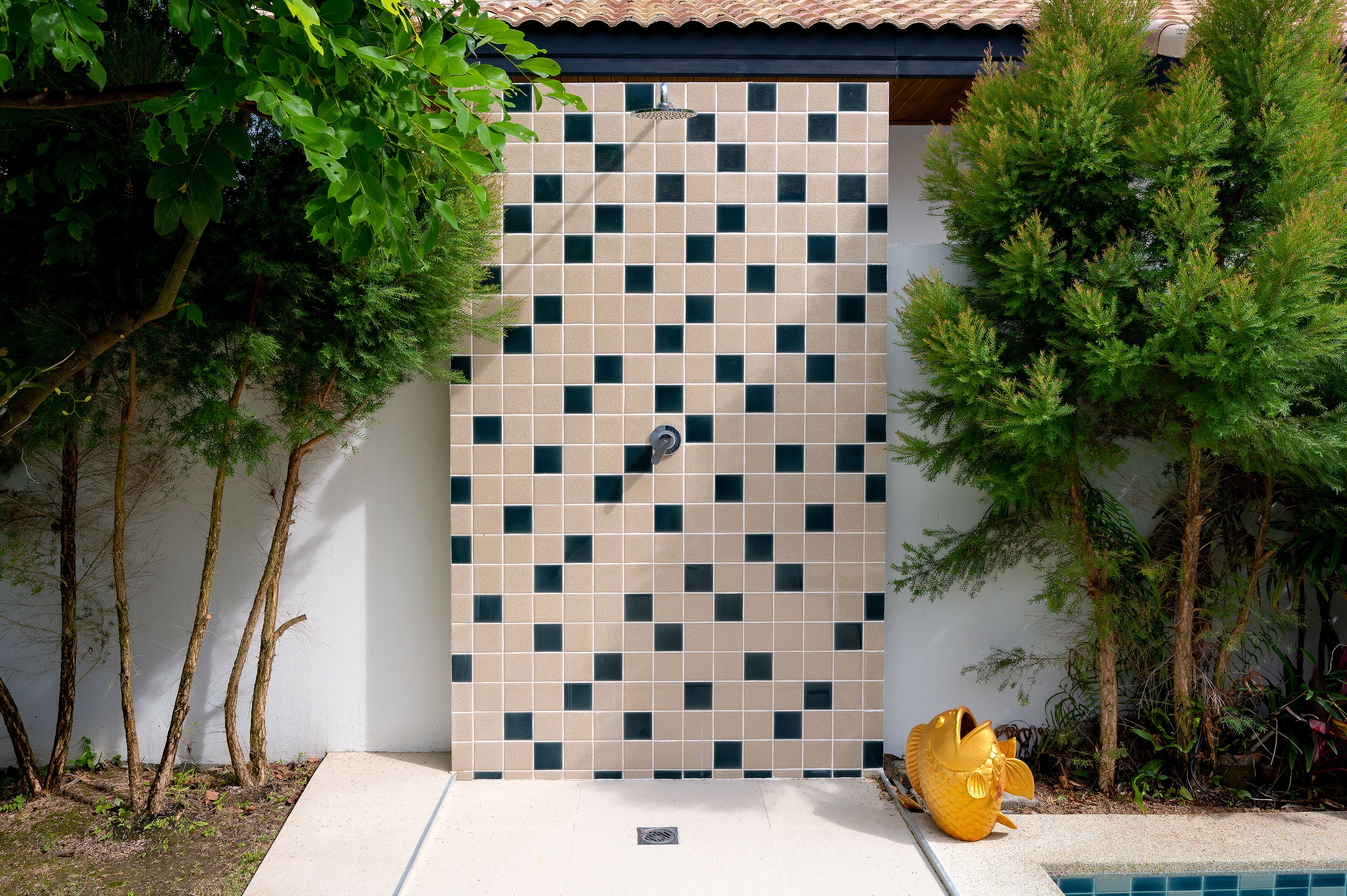 Pared de azulejos con zona de ducha, rodeada de árboles y una pequeña piscina con un objeto amarillo.
