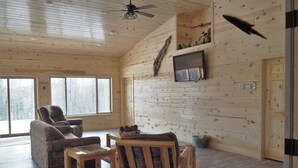A spacious room with wooden walls, a ceiling fan, a fireplace, and a large window offering a view of the outdoors. - Enjoy privacy in this newly built cabin with spectacular view of North Star Lake (Marcell)