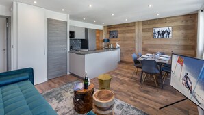 A modern living room with a sofa, a dining area with a table and chairs, a kitchen with a fridge and microwave, and a wooden wall with a picture.