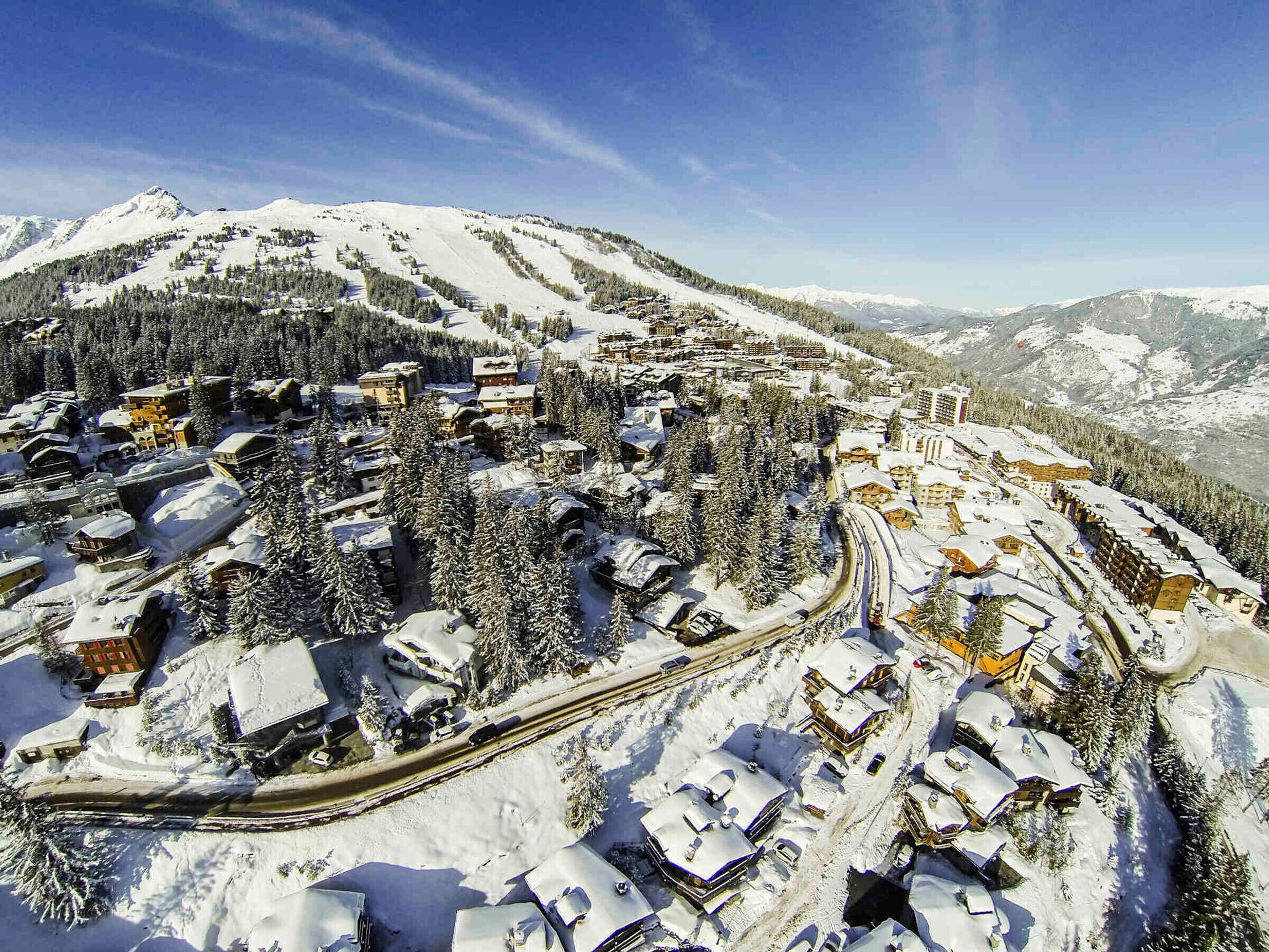 A snow-covered mountain town with buildings clustered around a winding road, surrounded by trees and mountains.