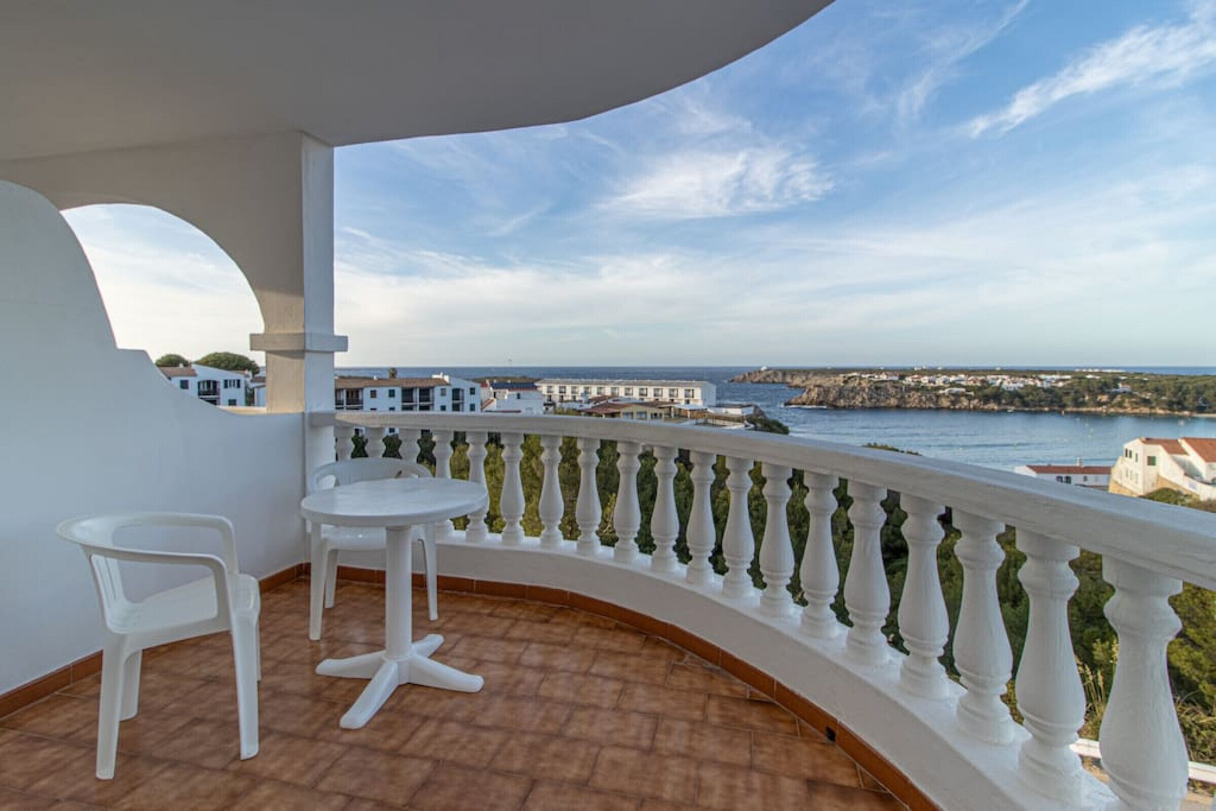 Un balcone con ringhiera bianca, un piccolo tavolo e due sedie. Il balcone si affaccia su una zona costiera con edifici e una distesa d'acqua.