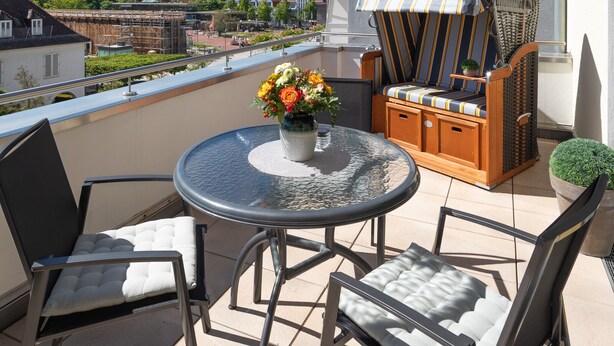 Un balcon avec une table ronde, deux chaises, un fauteuil en osier recouvert d’un tissu à rayures et un pot de fleurs.
