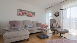 Un salon moderne avec un canapé, un pouf, une table basse et un fauteuil. Un tableau est accroché au mur et une lampe sur pied se trouve dans la pièce.