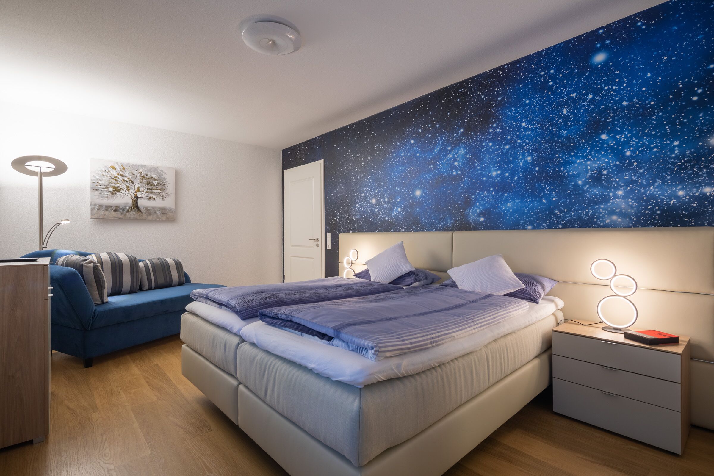 A bedroom with a bed, bedside table, nightstand, and a wall mural of a starry night sky.