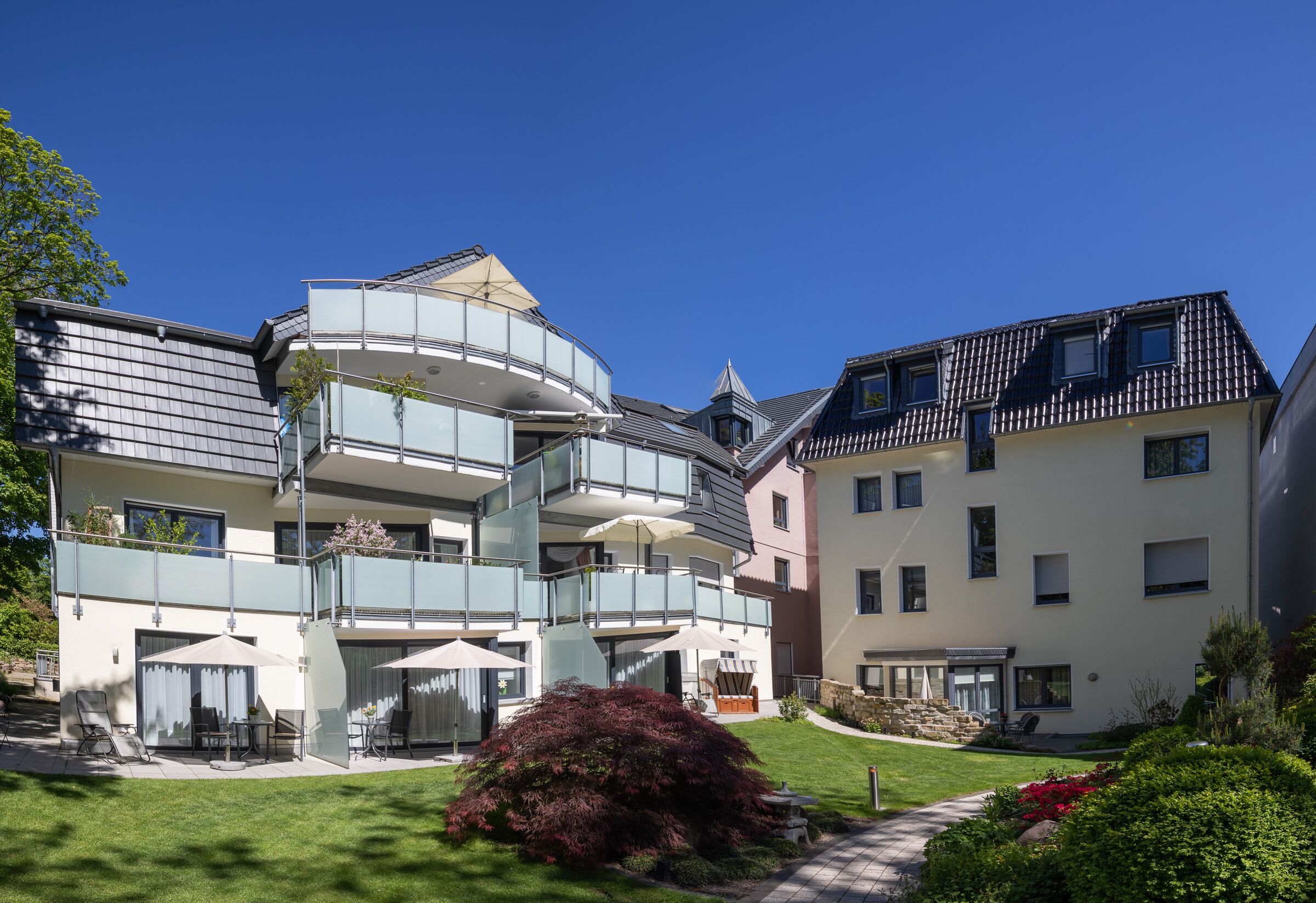 Un moderno edificio residencial de varios pisos con balcón de vidrio, zona de estar al aire libre y un jardín bien cuidado.