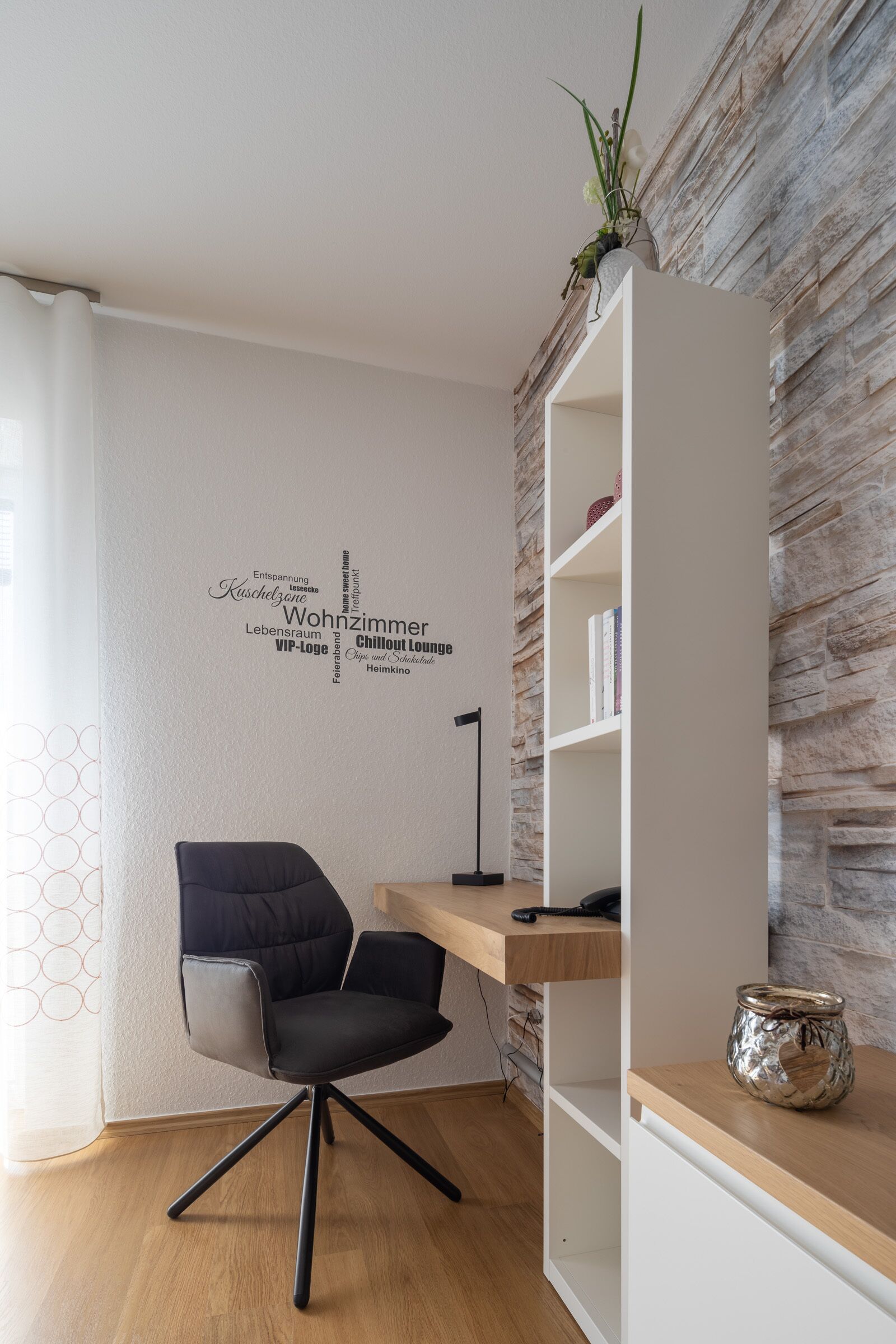 A modern room with a black office chair, a wooden desk, and a white shelving unit against a stone wall.