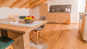 A dining area with a wooden table, chairs, and a bowl of fruit. There is a kitchen in the background with wooden cabinets and a sink. - Ferienwohnung (Lenggries)