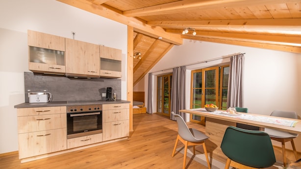 A modern kitchen with wooden cabinets and a built-in oven, a dining area with a table and chairs, and a large window with curtains. - Ferienwohnung (Lenggries)
