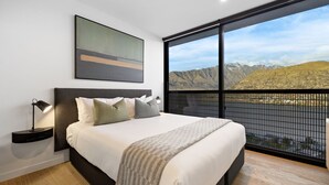 Quarto moderno com uma cama grande, mesa de cabeceira, luminária e vista para montanhas através de uma janela ampla.