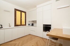 Une cuisine moderne avec des meubles blancs, un parquet en bois disposé en chevrons, une table en bois et une fenêtre avec un cadre en bois.