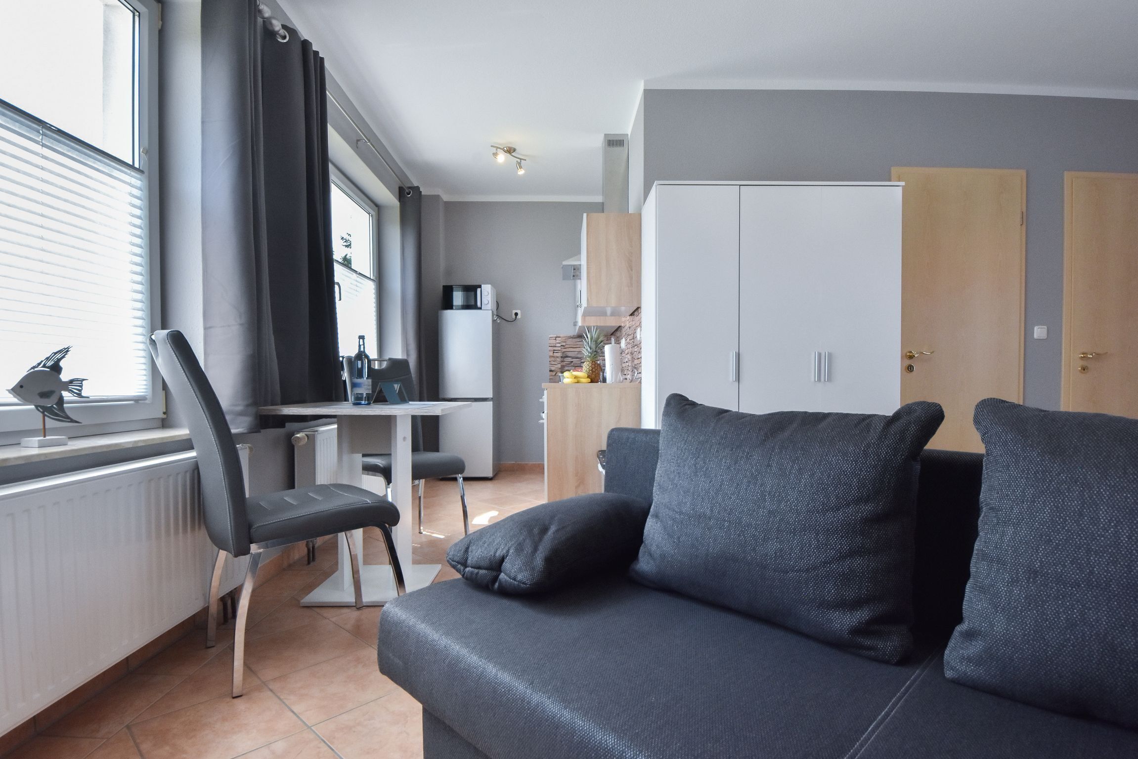 Ein modernes Wohnzimmer mit einem dunkelgrauen Sofa, einem Essbereich mit Tisch und Stühlen sowie einer K üche mit Schränken und einer Mikrowelle.