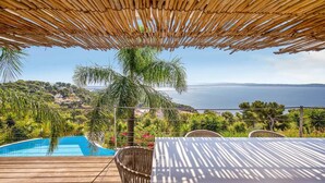 Zona piscina con tavolo e sedie, tetto di paglia e vista sul mare e sulle colline.
