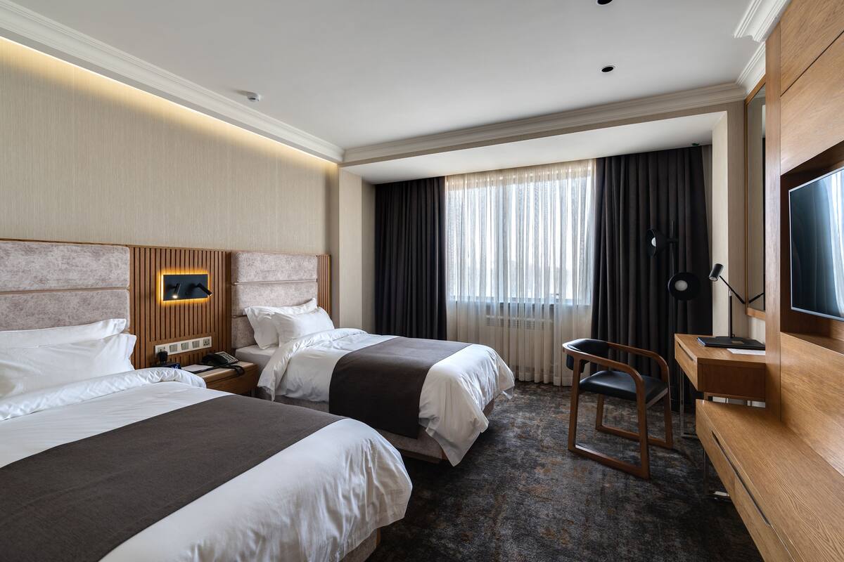 A hotel room with two beds, a desk, a chair, a TV, and a window with curtains.