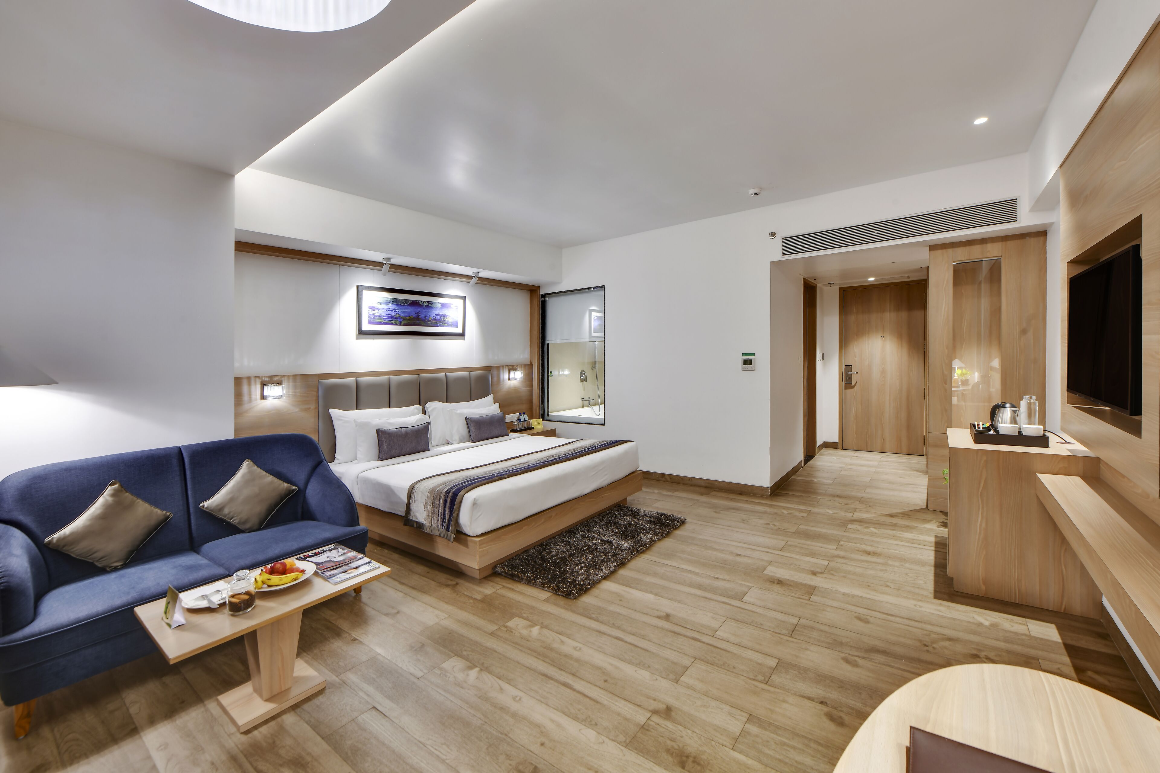 A modern hotel room with a bed, a sofa, a coffee table, a TV, and a wooden desk.