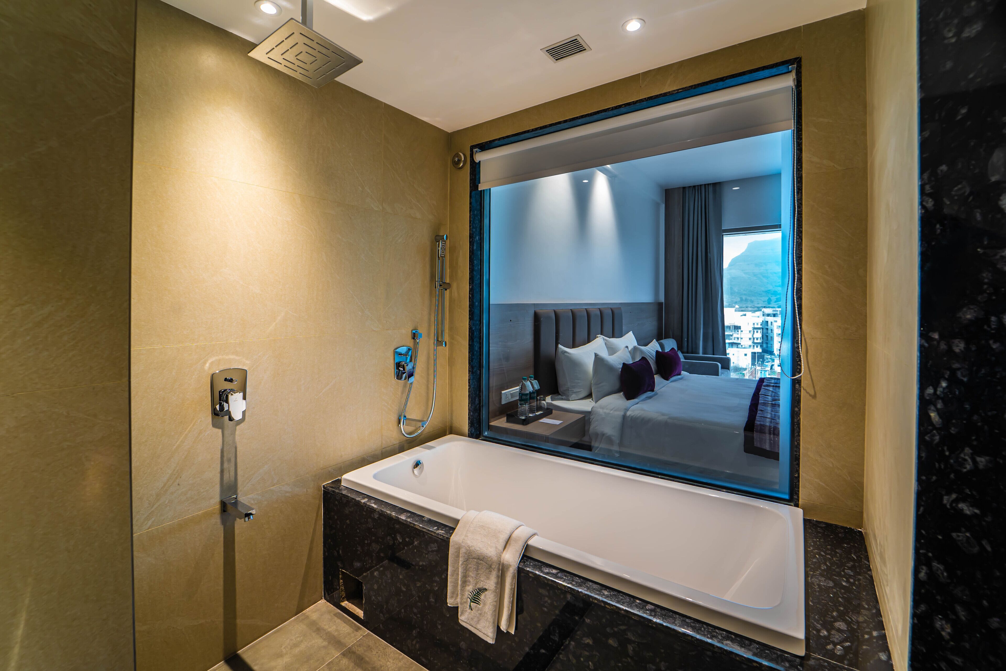 Een moderne badkamer met een groot bad, douche en een spiegel die een slaapkamer met bed en stadszicht weerspiegelt.